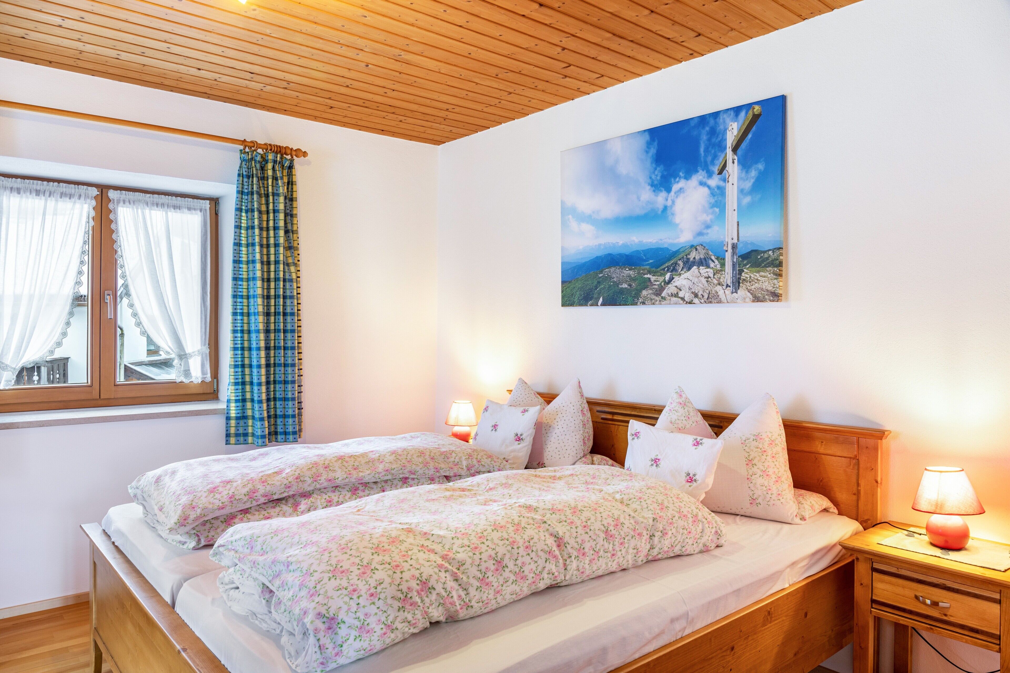 Una camera da letto con due letti, un soffitto in legno, una finestra con tende, un comodino con una lampada e un quadro appeso al muro.