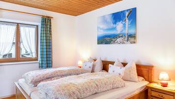 A bedroom with two beds, a wooden ceiling, a window with curtains, a nightstand with a lamp, and a painting on the wall.