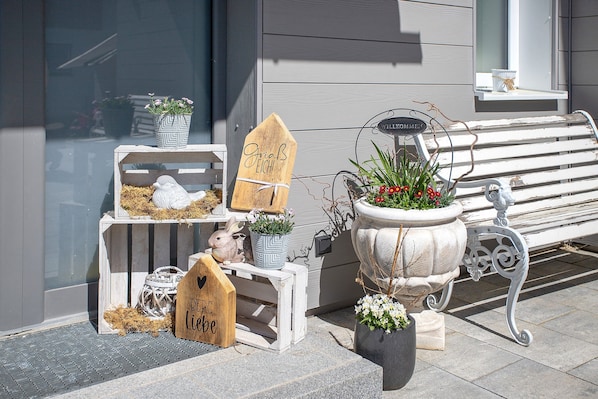 Un banc en bois, un jardinier avec des fleurs et une petite figurine d’oiseau blanc posée sur une marche.