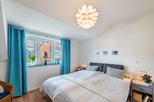 A bedroom with a bed, a window with curtains, a bedside table with a lamp, and a wall-mounted light fixture.