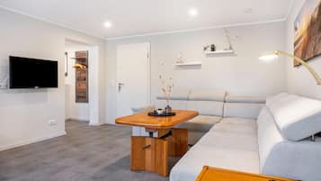 Een moderne woonkamer met een flatscreen televisie, een houten salontafel, een grijze bank en een wandplank met decoratieve objecten.