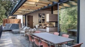 An outdoor dining area with a table and chairs, a wooden ceiling, and a view of the garden through large windows. - Riverside Haven Elegant House with Serene Forest (Avandaro)