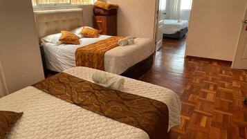 Una habitación de hotel con dos camas, un suelo de madera en patrón de espiga y una ventana con persianas.