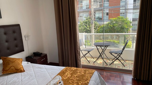 A hotel room with a bed, a nightstand, a window with a view of buildings and greenery, and a balcony with chairs and a table. - LIMA HOUSE MILANI (Lima)