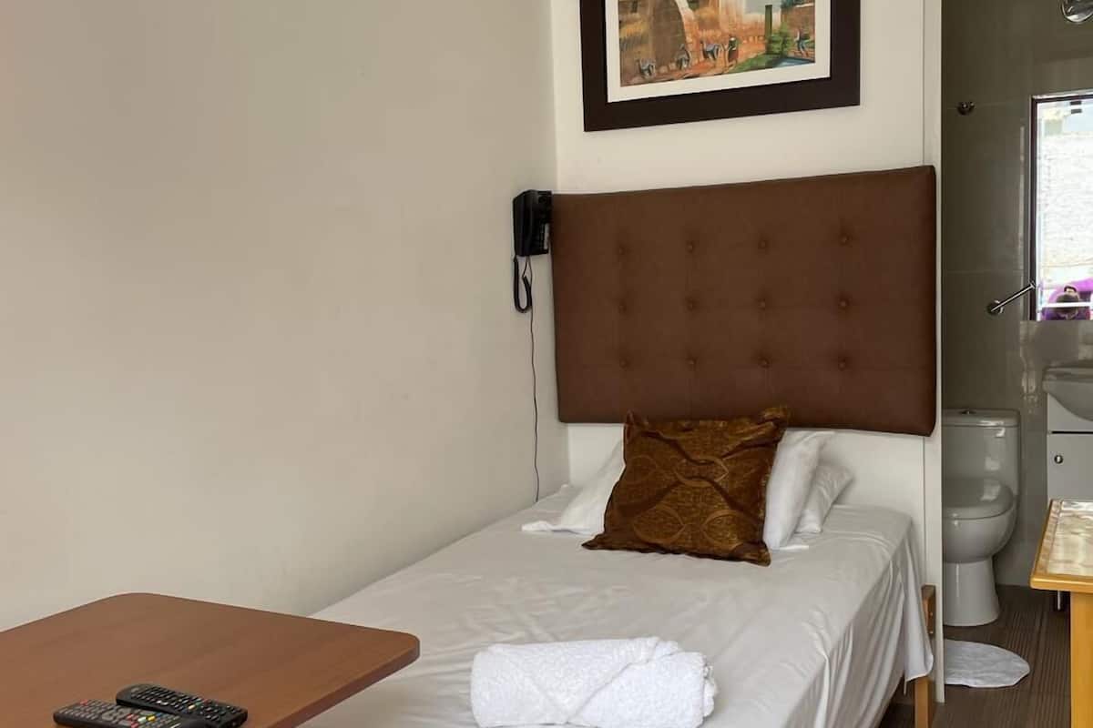 A hotel room with a single bed, a small table with a remote control, a telephone, and a bathroom visible through an open door.