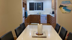 A dining area with a long white table, six black chairs, and a central paper towel dispenser. A kitchen is visible in the background with wooden cabinets and a window.