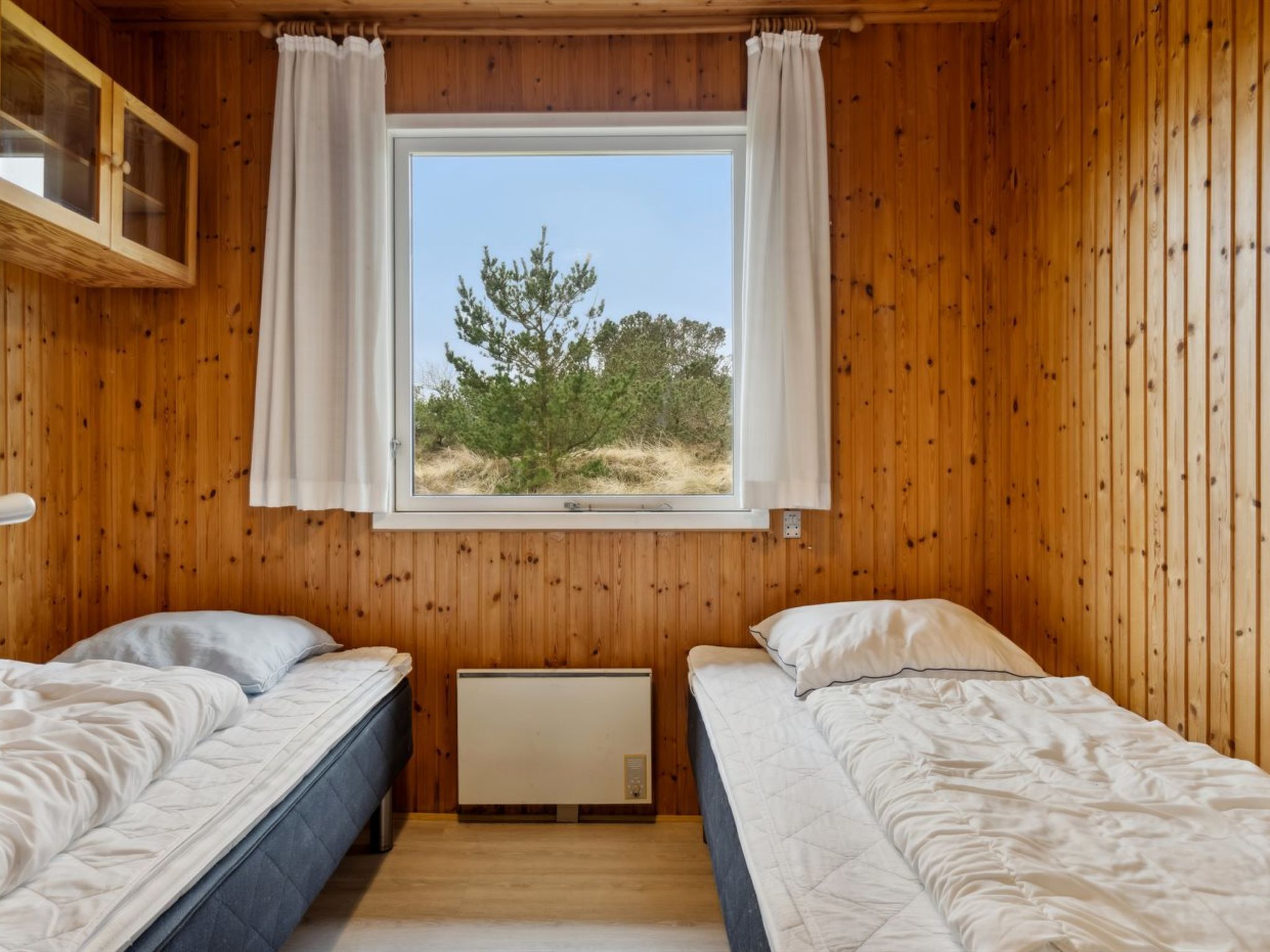 A room with wooden walls, two beds, a window with curtains, and a radiator.