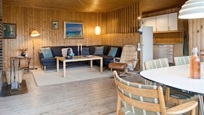 En mysig stuga med träpanel i taket, ett vardagsrum med soffa och soffbord, ett matbord med stolar och ett köksutrymme.