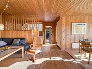 Ein modernes Interieur mit Holzbalkendecke, einer Couch, einem Sessel, einem Esstisch und Blick durch eine offene Tür.
