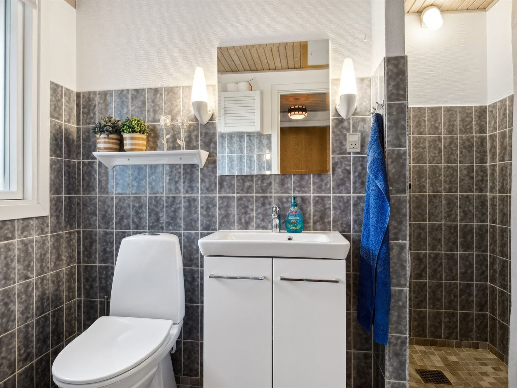A bathroom with a toilet, sink, and shower area. There are wall-mounted lights, a shelf with plants, and a towel hanging on the wall.