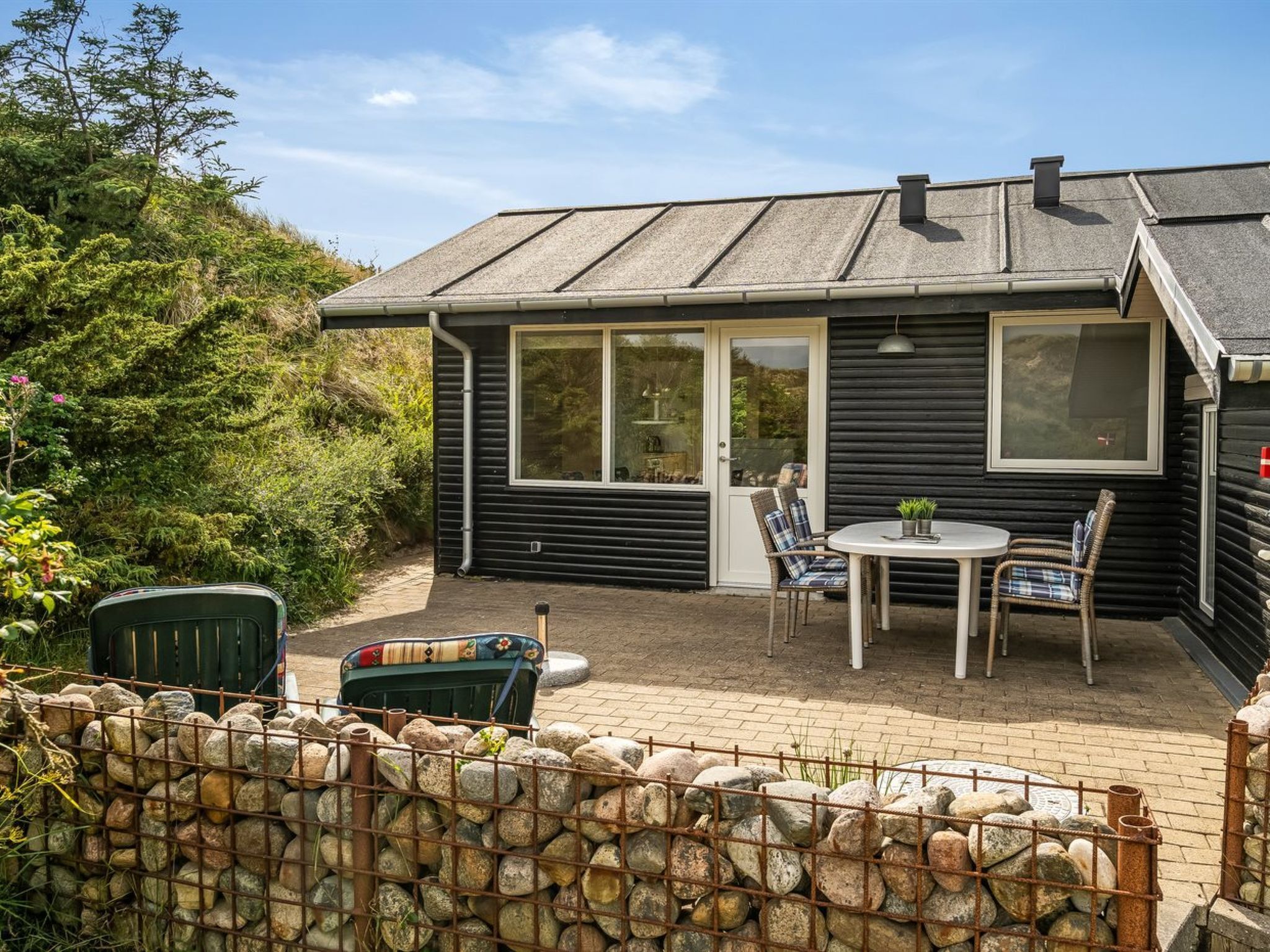 A black wooden house with a patio area, including a table and chairs, surrounded by a stone wall.