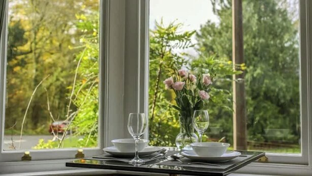 A dining table set with white plates, glasses, and a vase of pink flowers, positioned by a large window with a view of greenery.