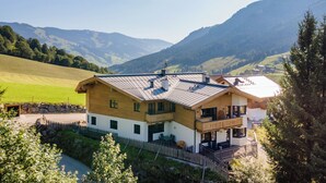 A two-story wooden house with a sloped roof, surrounded by green fields and mountains.