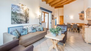 A cozy living room with a wooden ceiling, a sofa with patterned cushions, a dining table with chairs, and a kitchen area with wooden cabinets.