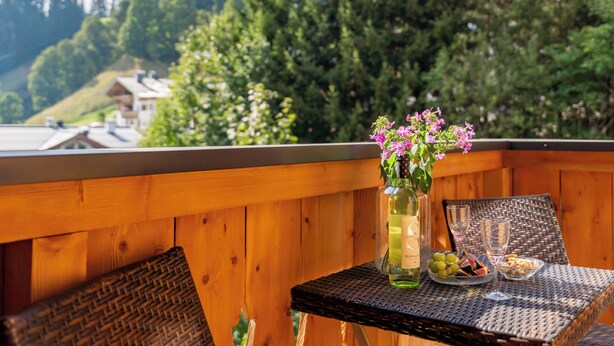 A wooden balcony with a wicker table set for two, a bottle of wine, and a vase with flowers.