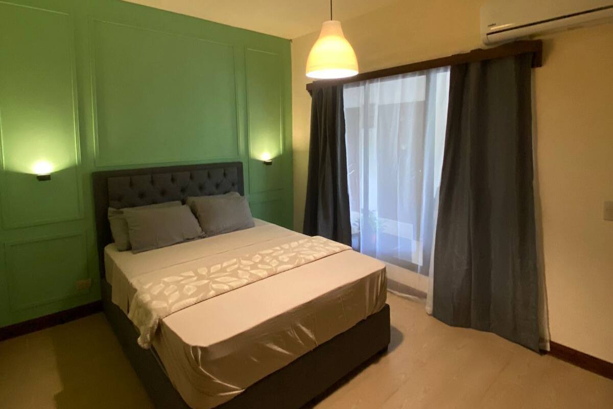 A bedroom with a bed, a hanging light fixture, a window with curtains, and an air conditioning unit.