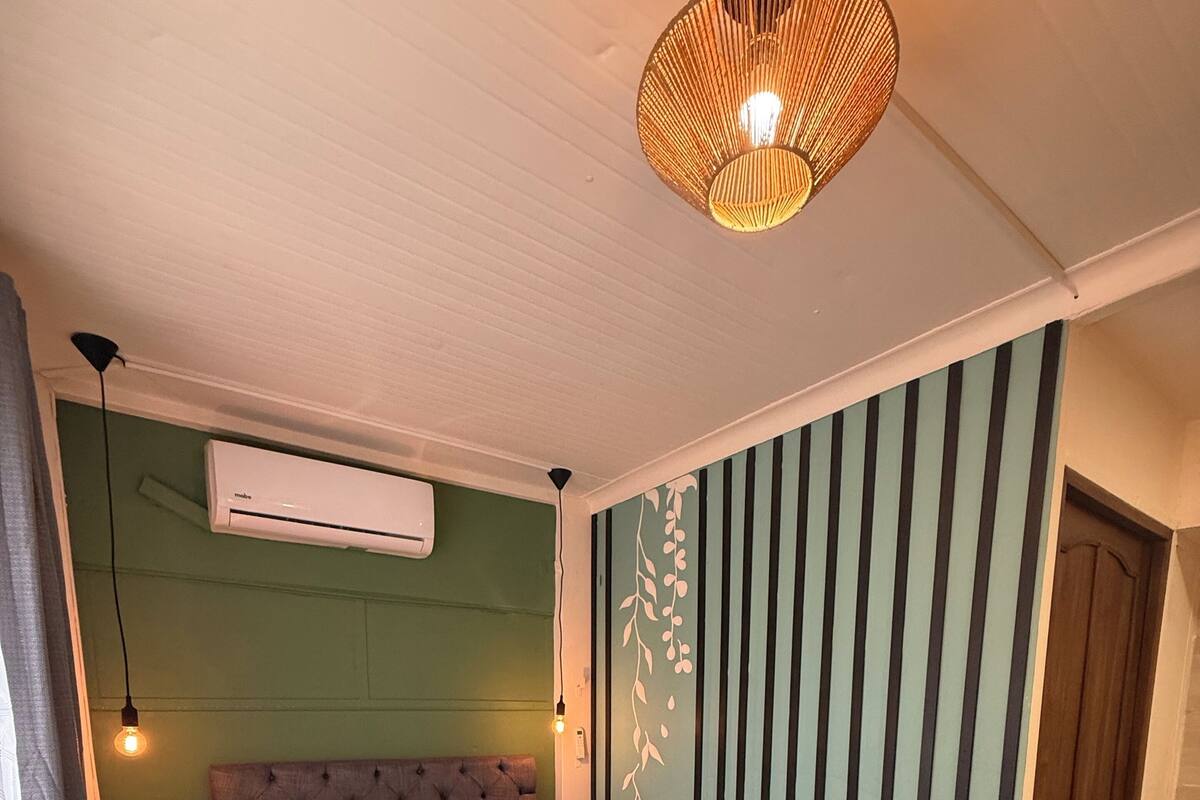 A bedroom with a large bed, a wall-mounted air conditioner, a hanging light fixture, and a striped wall decoration.