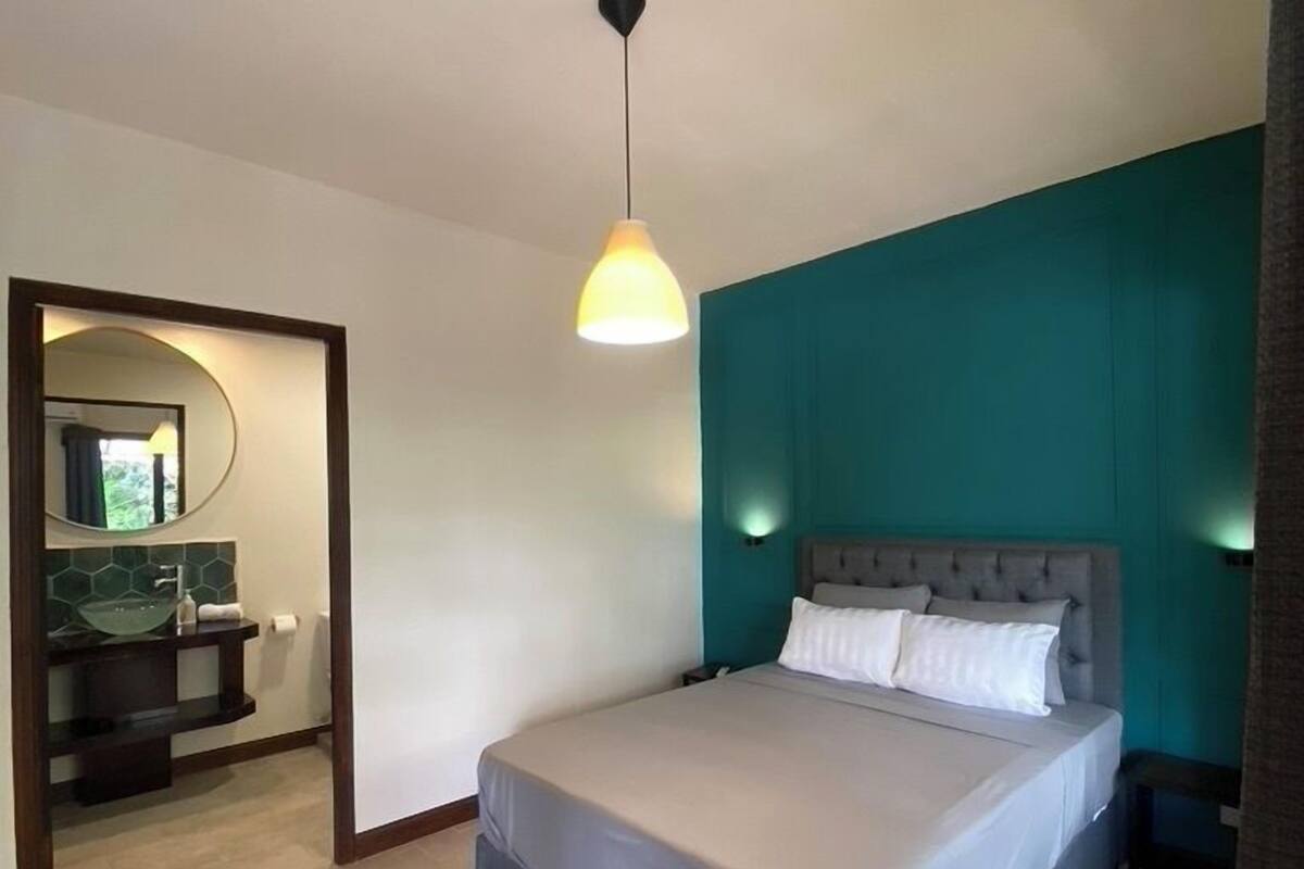 A bedroom with a bed, a nightstand, a bedside lamp, a mirror, and a bathroom with a sink and mirror.