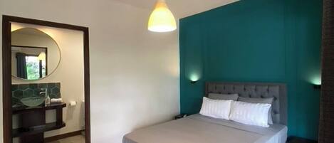 Un dormitorio con cama, mesita de noche, lámpara de noche, espejo y baño con lavamanos y espejo.
