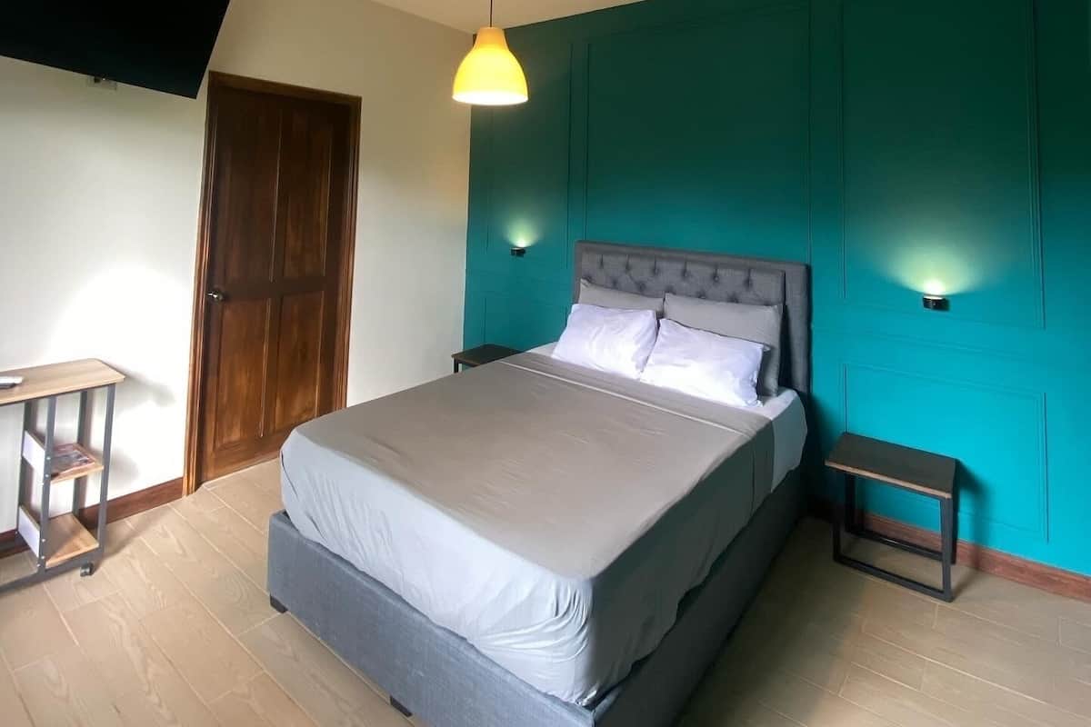A bedroom with a bed, a wooden door, a TV mounted on the wall, and a small table with a chair.