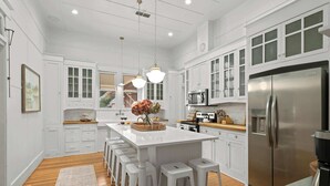 A spacious kitchen with white cabinetry, a central island with stools, a stainless steel refrigerator, and a hanging light fixture.