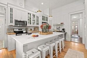 Uma cozinha com armários brancos, uma ilha grande com banquetas, um micro-ondas e um lustre.