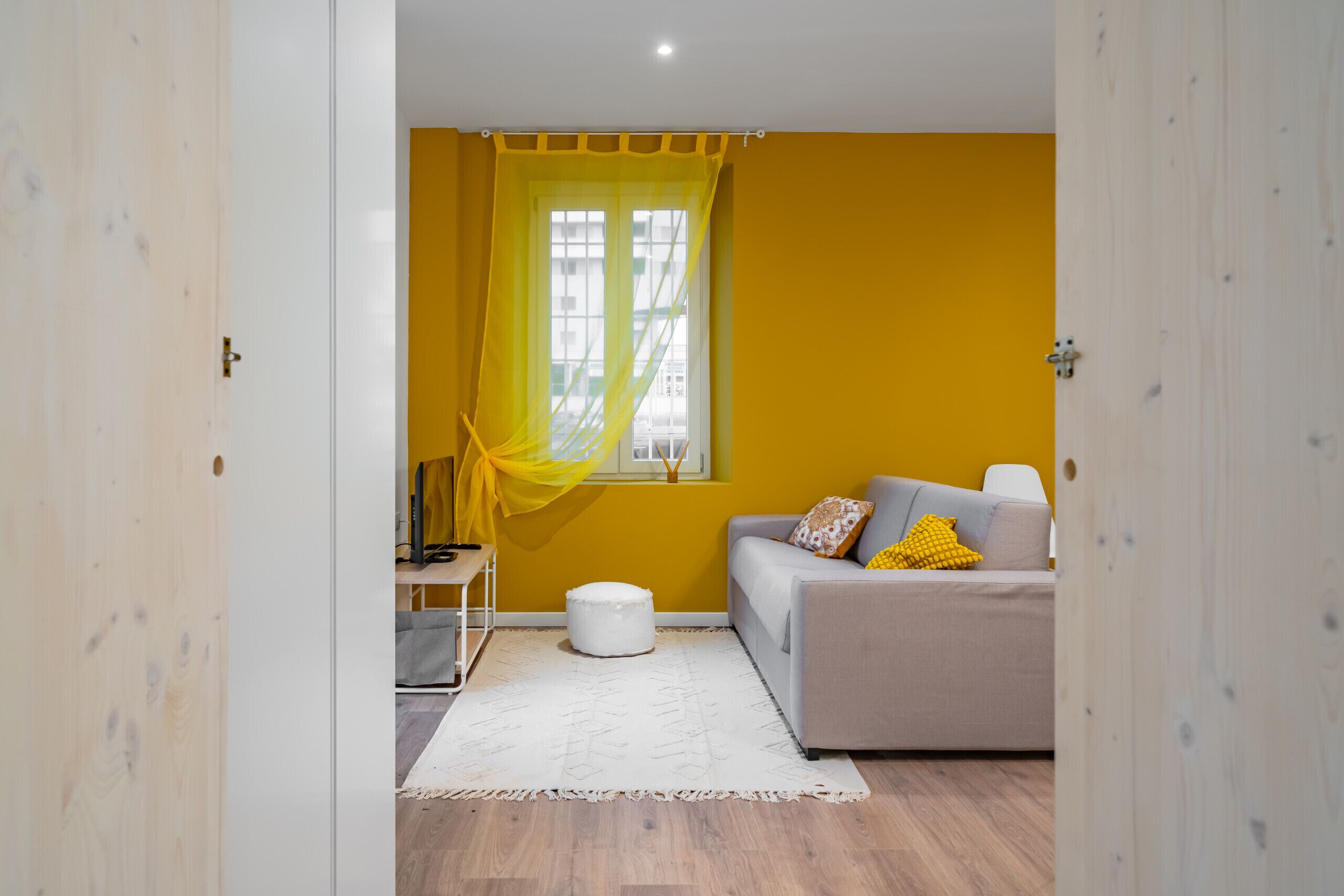 A room with a sofa, a window with yellow curtains, a white ottoman, and a white rug.
