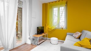 A modern living room with a sofa, a TV on a stand, a window with yellow curtains, and a white door with a view of the outdoors. - Cascina Conicchio 1 (Brescia)
