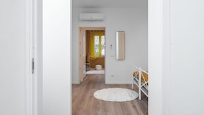 A hallway with a white door, a white wall, a white floor, a white bed with a yellow blanket, and a white rug. - Cascina Conicchio 1 (Brescia)