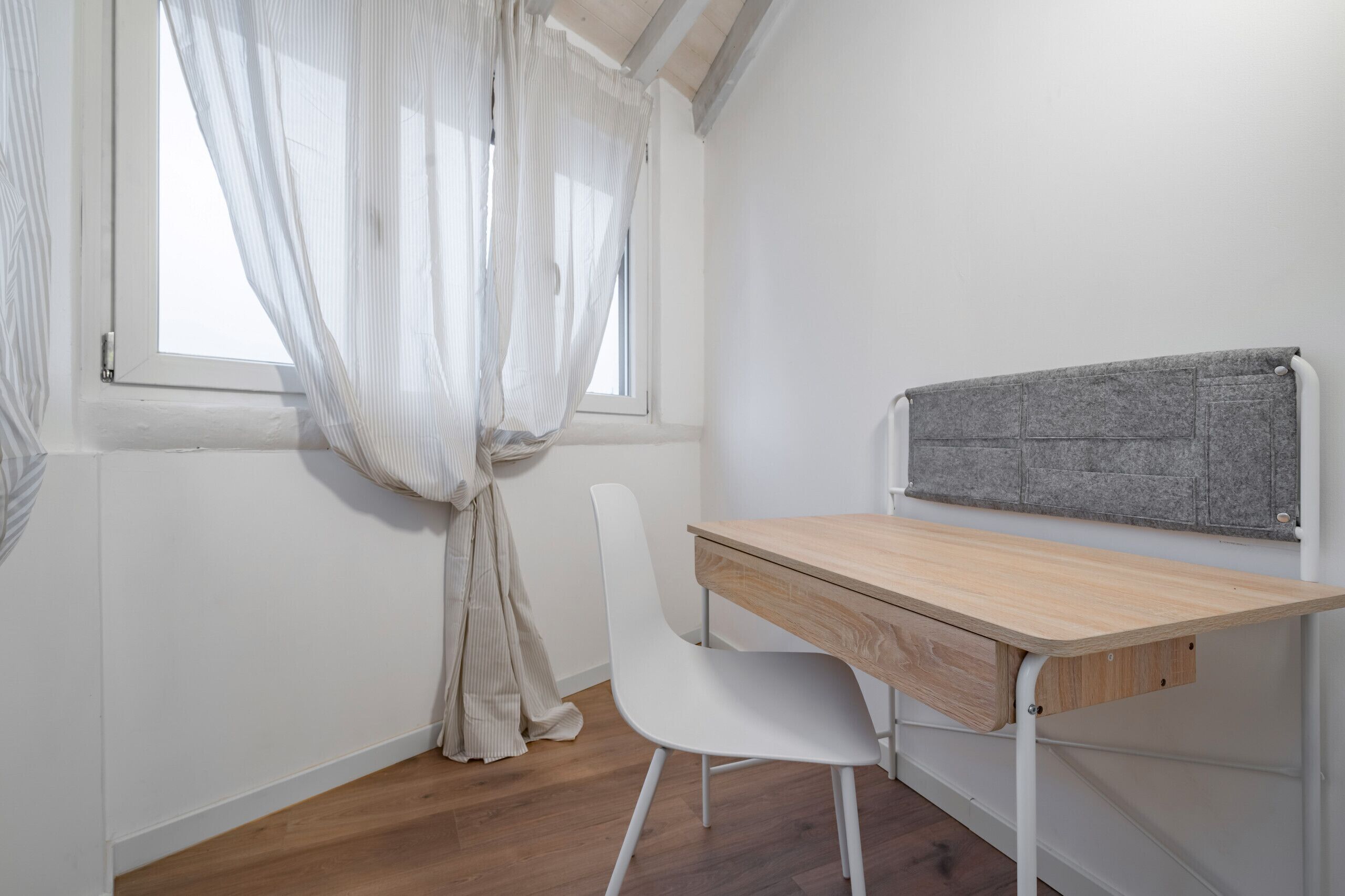 A small, neatly organized room with a wooden desk, a white chair, and a grey headboard.