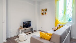 Moderni olohuone, jossa on valkoinen, litteänäyttöinen televisio, harmaa sohva, jossa on keltaiset koristetyynyt, valkoinen rahi ja ikkuna, jossa on keltaiset verhot.