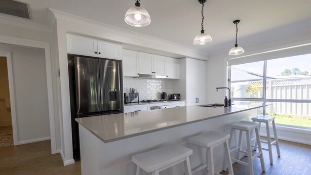 A modern kitchen with white cabinetry, a stainless steel refrigerator, a large island with bar stools, and pendant lighting. - Luxury on Lesmurdie - Close to Zoo & Golf Club (Dubbo)