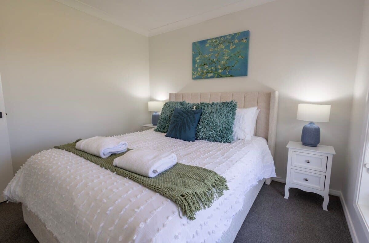 A neatly made bed with white and green linens, a blue pillow, and a green throw blanket. There is a painting on the wall above the bed, and two bedside tables with lamps.