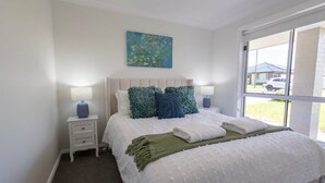 A neatly made bed with a white and blue color scheme, a small bedside table with a lamp, and a window with a view of the outside. - Luxury on Lesmurdie - Close to Zoo & Golf Club (Dubbo)