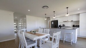 A modern kitchen with white cabinetry, a central island, pendant lighting, and a dining area with white chairs and a table. - Luxury on Lesmurdie - Close to Zoo & Golf Club (Dubbo)