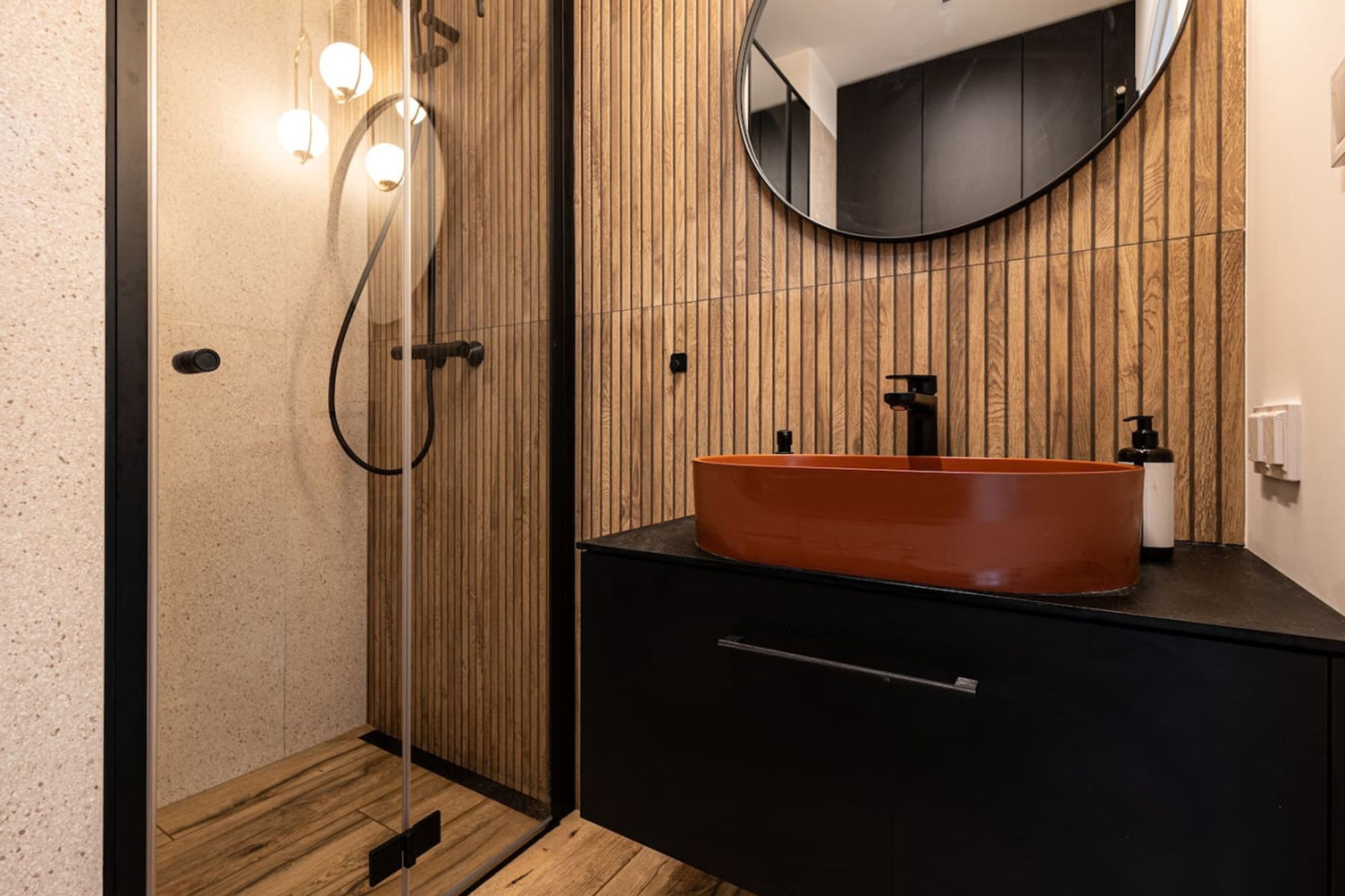 A modern bathroom with a wooden panel wall, a round mirror, a black sink with an orange basin, and a glass shower enclosure.