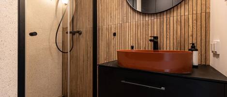 A modern bathroom with a wooden panel wall, a round mirror, a black sink with an orange basin, and a glass shower enclosure.