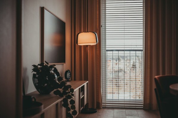 A modern hotel room with a large window, a flat-screen TV mounted on the wall, a small table with a potted plant, and a floor lamp.