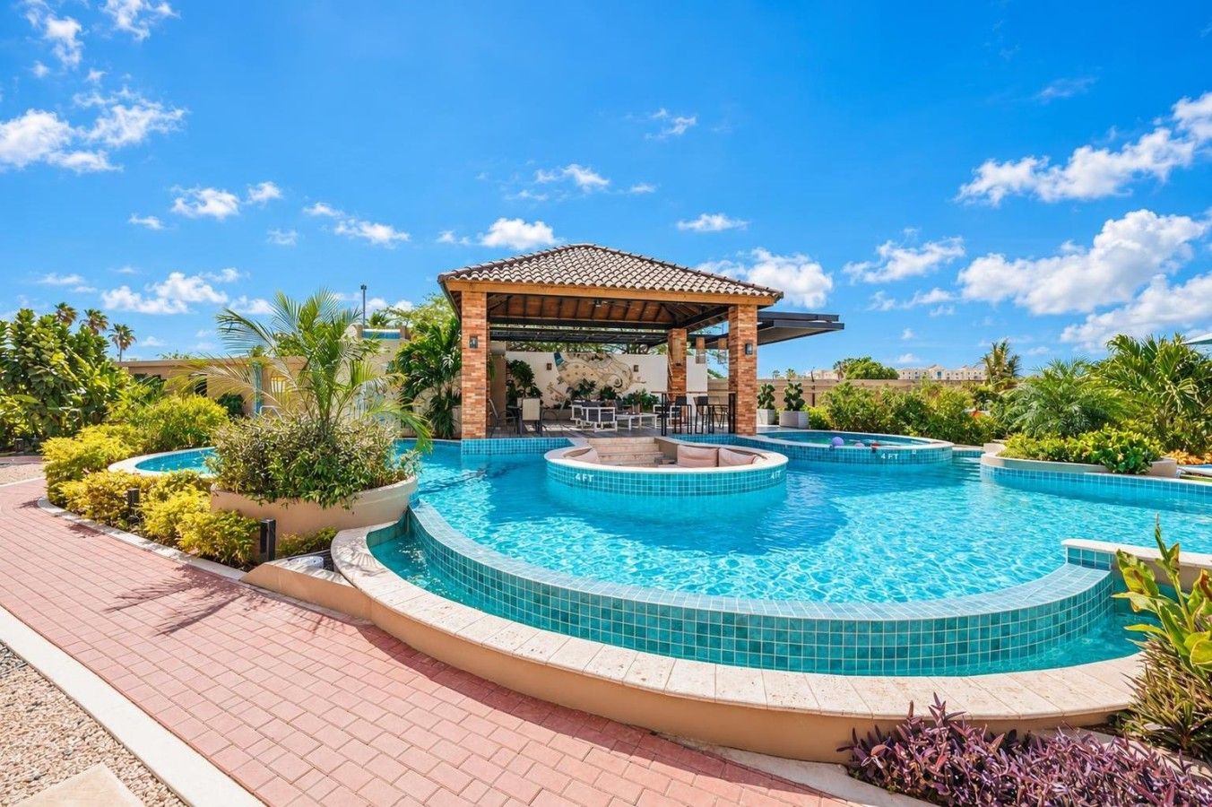 A swimming pool with a sunken hot tub, surrounded by lush greenery and a covered seating area.