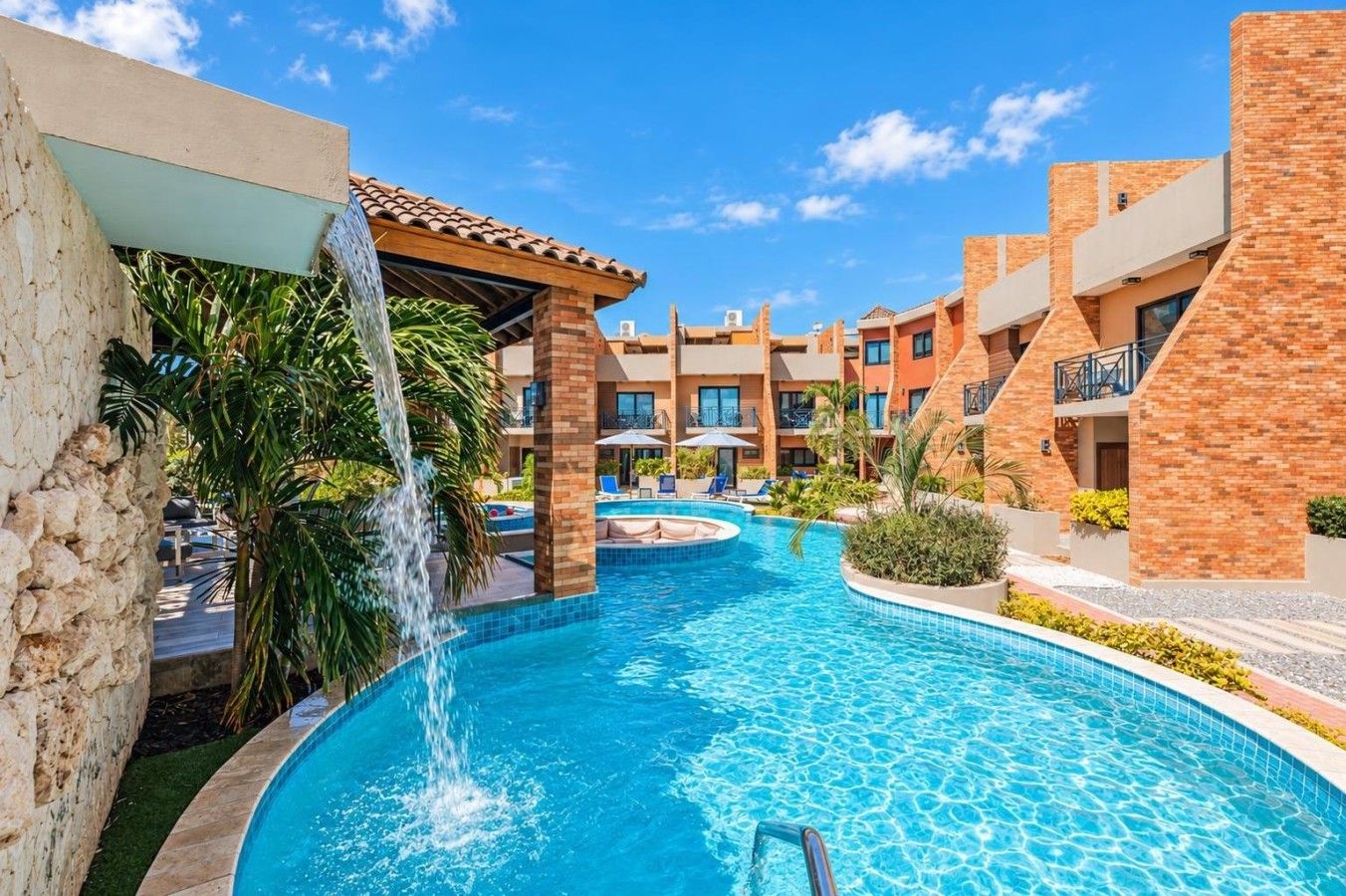 A swimming pool with a waterfall feature, surrounded by multi-story buildings with balconies and outdoor seating.