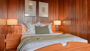 A wooden-paneled bedroom with a bed, two bedside lamps, a nightstand, and two framed botanical prints on the wall. - Chalet do Relógio d’Água, a Home in Madeira (Ponta do Sol)