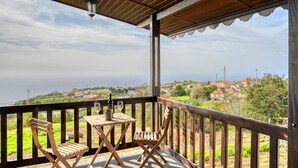 Un balcon en bois, agrémenté d’une table et de chaises, qui offre une vue sur un paysage côtier avec des maisons et de la végétation.