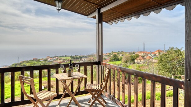 A wooden balcony with a table and chairs, overlooking a coastal landscape with houses and greenery. - Chalet do Relógio d’Água, a Home in Madeira (Ponta do Sol)