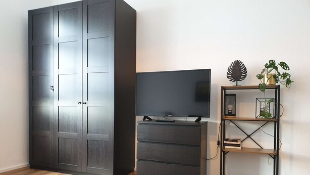 A room with a large wardrobe, a flat-screen TV on a wooden stand, and a plant on a metal shelf. - Luxuriöse großzügige Wohnung mit 3 Schlafzimmer, Terrasse (Solingen)