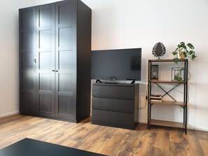 Quarto com um guarda-roupa espaçoso, uma televisão de tela plana sobre um suporte de madeira, e uma planta em uma estante de metal.