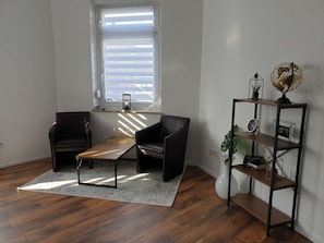 A room with a wooden floor, two leather chairs, a small table, a bookshelf, and a window with blinds.