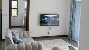 Un salon au design moderne, comprenant un ensemble de canapés gris, un téléviseur à écran plat fixé au mur et un plafond avec des poutres apparentes.