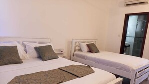 Une chambre avec deux lits simples, chacun doté de draps blancs et d’un couvre-lit marron à motifs. On trouve un banc blanc devant les lits, et un appareil de climatisation fixé au mur.