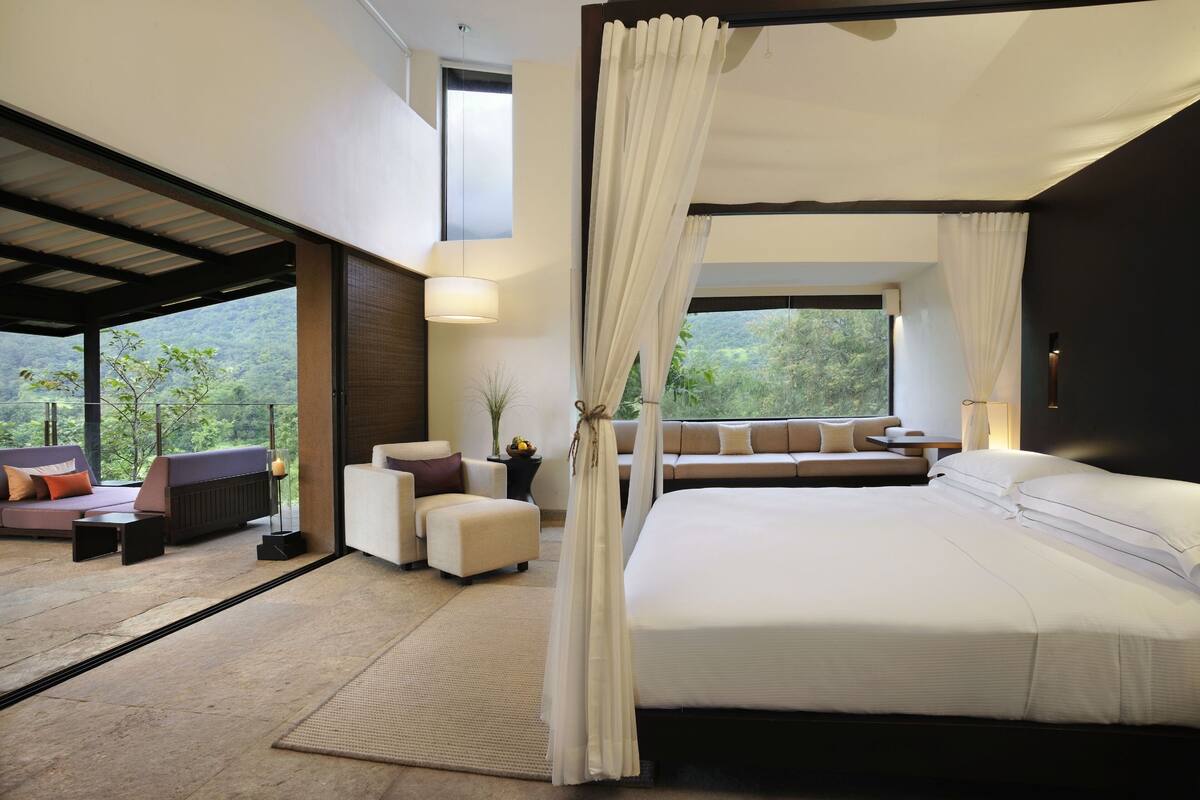 A modern bedroom with a large bed, a sofa, and a view of a lush landscape.