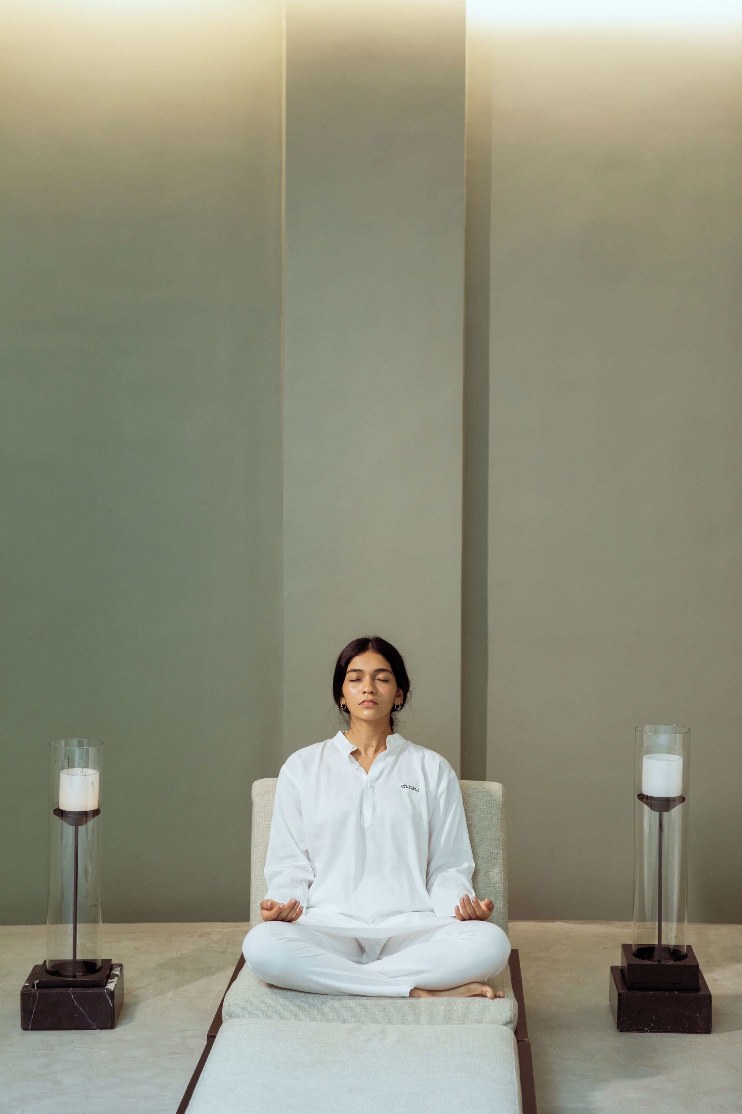A person meditating in a minimalist room with a plain wall, a cushioned bench, and two candle stands.